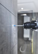 Eine Bohrmaschine bohrt ein Loch in eine Fliesenwand; der Bohrstaub ist sichtbar.