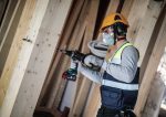 Ein Arbeiter mit Sicherheitsausrüstung bohrt auf einer Baustelle in einen Holzbalken.